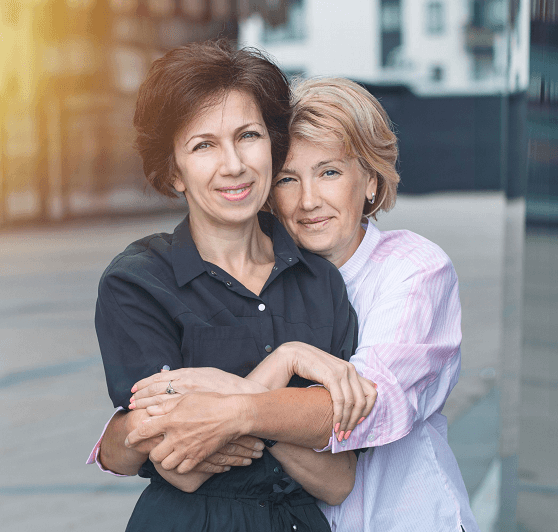 An older lesbian woman embraces her lover from behind, both smiling at camera. 