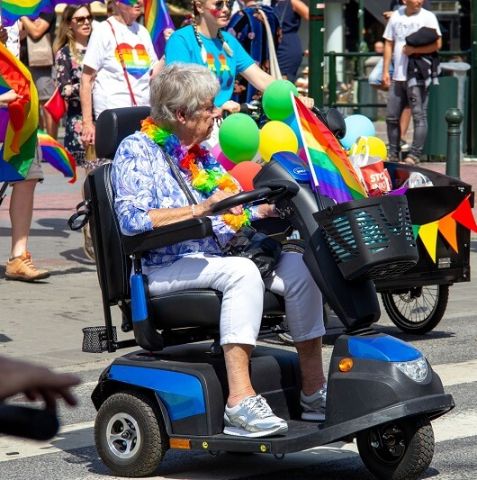 An older woman in a motorized mobility device wearing rainbow flowers in a pride parade.