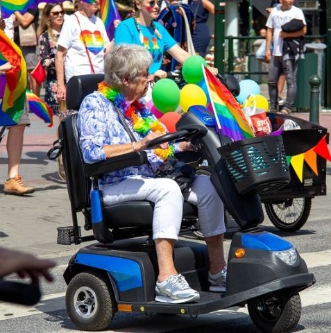 An older woman in a motorized mobility device wearing rainbow flowers in a pride parade. 